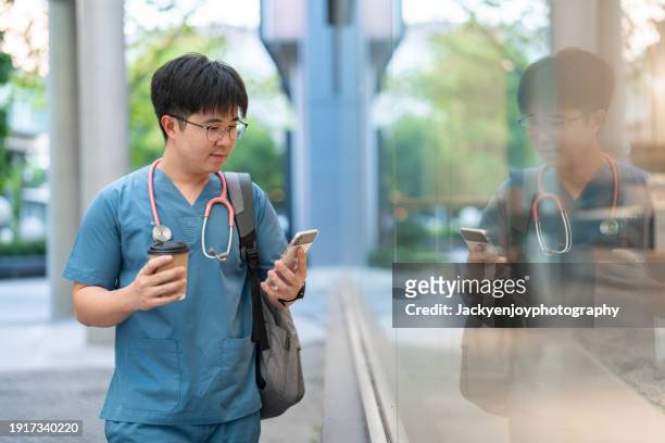 during his break from work, a young asian male healthcare worker is seen sipping hot coffee and holding a cell phone. - hot nurse stock-fotos und bilder