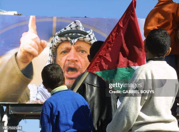 Huge banner of Palestinian leader Yasser Arafat making a speech adorns the street as a boy holding the Palestinian flag peers around the shoulders of...