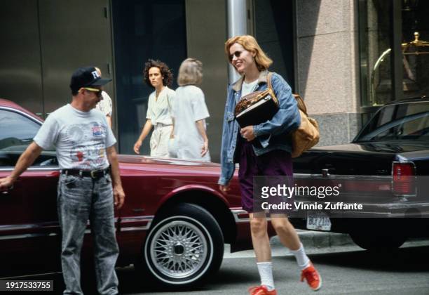 American actress Geena Davis, wearing a denim jacket and purple shorts, carrying a ringer binder and a baseball mitt with the strap of a shoulder bag...