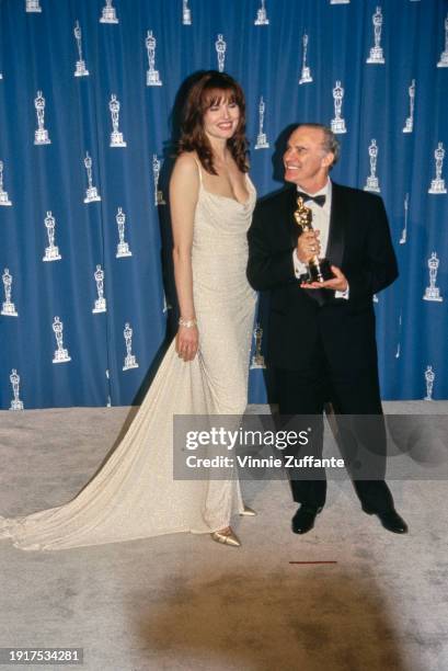 American actress Geena Davis, wearing a white evening gown, with American film editor Michael Kahn, who wears a tuxedo and bow tie, in the press room...