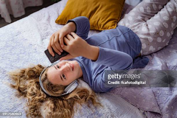 happy teenager girl laying on her bedroom using headphones and searching music in her smartphone - kid headphones stock pictures, royalty-free photos & images