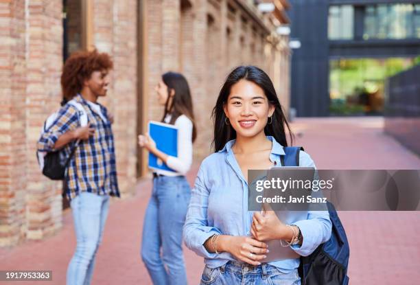 retrato de una joven estudiante asiática mirando a la cámara sonriendo. al fondo, dos estudiantes hablando en el campus - edificio público fotografías e imágenes de stock