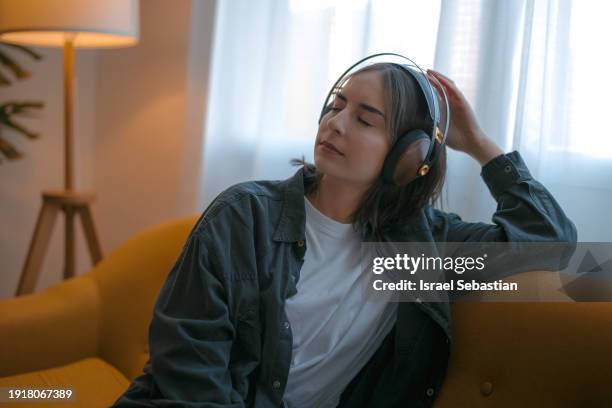 young woman with closed eyes relaxing on sofa and listening to music on headphones. - fashion suit stock pictures, royalty-free photos & images