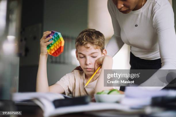 mother assisting son holding push pop toy while doing studies at home - neurodiversidad fotografías e imágenes de stock