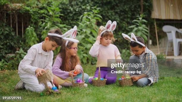 group of children sitting on grass and opening easter eggs on easter egg hunt game in back yard garden - easter egg hunt stock pictures, royalty-free photos & images