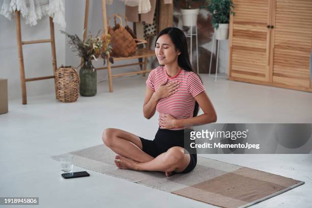 happy peaceful asian woman practicing diaphragmatic breathing, doing yoga at home, sitting in lotus position with eyes closed holding hands on chest and stomach, meditating in morning - woman flat chest 個照片及圖片檔