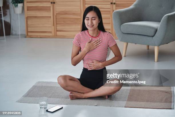 asian woman practicing breathing exercises while doing yoga in morning, sitting in lotus position with eyes closed holding hands on chest and stomach, meditating at home. mindfullness and mental health - woman flat chest 個照片及�圖片檔