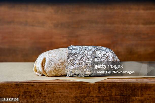 burrito partially wrapped in aluminum foil on wood table - burrito stock pictures, royalty-free photos & images