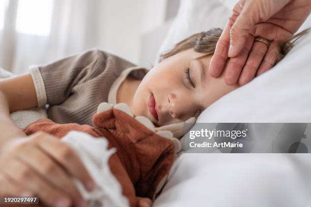 sick child sleeping at home in bed with flu, cold, covid, bronchitis or pneumonia, virus or infection and his mother's hand touching his forehead checking temperature - koorts stockfoto's en -beelden