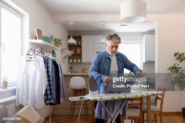 hombre maduro planchando ropa en casa - un solo hombre mayor camisa fotografías e imágenes de stock