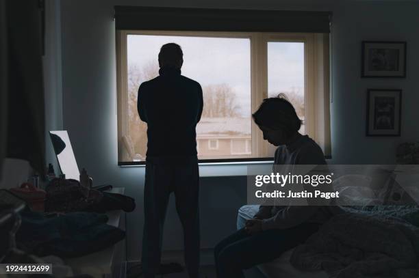 a silhouetted couple are in their bedroom. the woman sits on the bed deep in thought. the man looks out of the window with his arms crossed - negative emotion - menselijke relaties stockfoto's en -beelden
