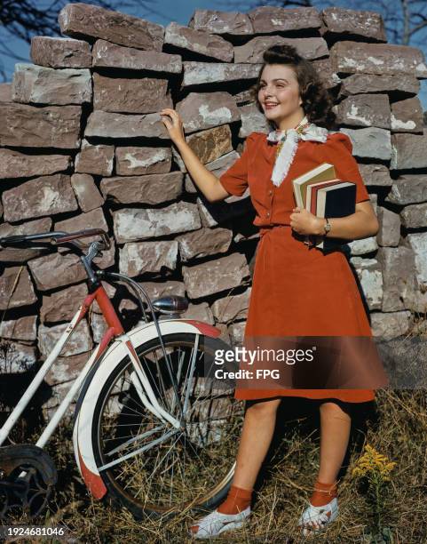 Woman holding books and near a bike stands next to a stone wall, US, circa 1996.