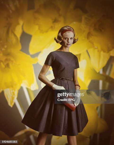 Fashion model wearing brown short sleeve flared dress with bow belt straight neckline, cream-colored gloves and leather clutch, on a daffodil...