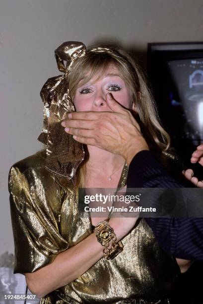 American actress Patti D'Arbanville wearing a gold outfit with a gold bow in her hair, an unspecified man's hand over her mouth, partially obscuring...