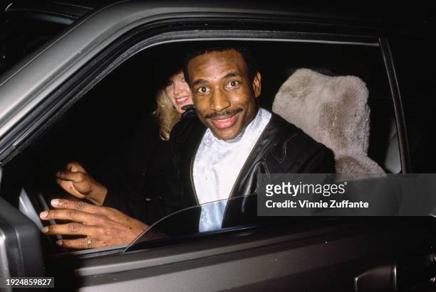 American football player Eric Dickerson wearing a black jacket with black leather trim over a white shirt, with a blonde woman sitting in the...