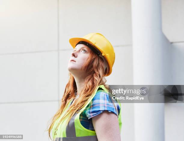 young woman in hardhat and hi-viz bib looks up seriously in front of a building - waistcoat stock pictures, royalty-free photos & images