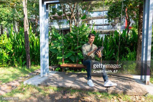 latin american businessman reading the articles using his digital tablet for further discussing with his team about business trend during sitting on a wooden swing at the park. - further stock pictures, royalty-free photos & images