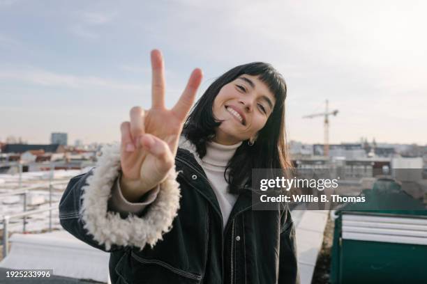 portrait of woman giving peace sign while standing on rooftop - peace sign gesture stock pictures, royalty-free photos & images