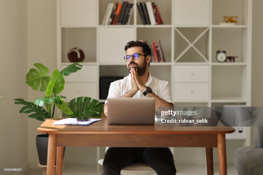 Serious pensive thoughtful young businessman or entrepreneur in modern contemporary office looking at and working with laptop and paper documents making serious and important business decision