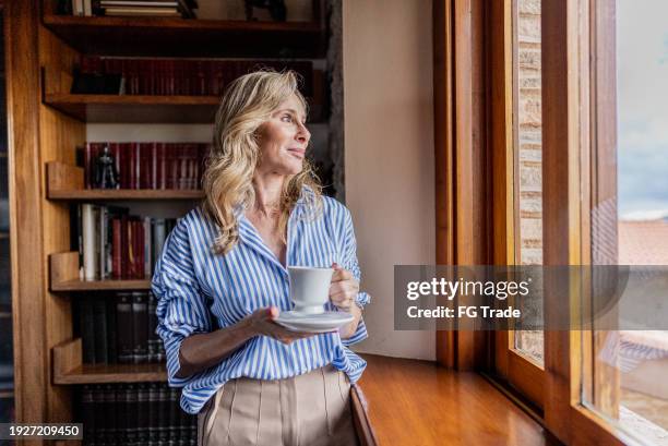 femme mûre contemplant tout en regardant par la fenêtre à la maison - richesse photos et images de collection