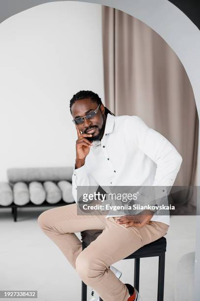 full body portrait of pensive young african man sitting on the chair at living room. - portrait of pensive young businessman wearing glasses stock-fotos und bilder