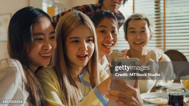 group of young asian people watching smartphone and having fun sitting at dining table at home. multicultural friends enjoying spending together college house party. - virtual thanksgiving stock pictures, royalty-free photos & images