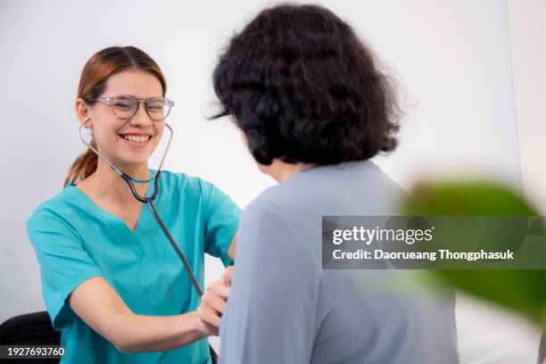 young nurse or caregiver sitting on sofa holding stethoscope listen heartbeat with senior woman in living room at home, doctor check cardiologist disease with elderly, medical and insurance. - gerontology stock pictures, royalty-free photos & images