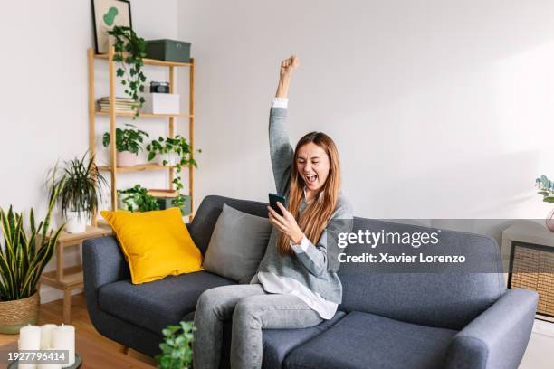 happy young adult woman winning money on smartphone app. young lucky female feeling excited after reading unexpected online message on smartphone. - good news bad news stock pictures, royalty-free photos & images