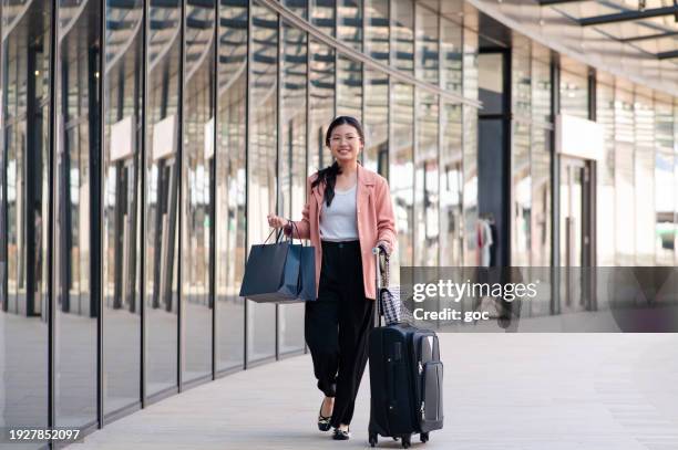 a young beautiful asian business woman with shopping bags in hand while pulling wheeled suitcases at airport terminal - financial freedom stock pictures, royalty-free photos & images