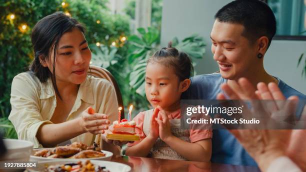 asian family blowing birthday candle on a cake while celebrating and having fun sitting at dining table at backyard outside home. multi-generation family enjoying spending together. - parents children blow candles asians foto e immagini stock