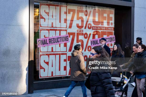 People out shopping on Oxford Street walk past large scale January sale signs in red and white for major high street clothing retail shops on 15th...