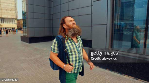 bearded red-haired man looking up walking along a gray building - senior man grey long hair stock pictures, royalty-free photos & images