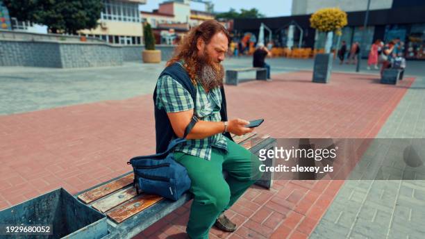 redhead bearded man with long hair reading message on a phone sitting on a city bench - senior man grey long hair stock pictures, royalty-free photos & images