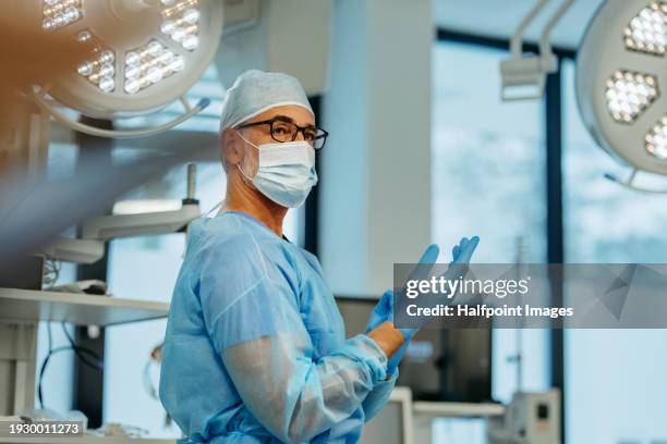 doctor putting on surgical gloves in operating room. surgeon dressed in surgical attire - head cover or surgical cap, scrub suit and surgical mask. obstetrician preparing for the delivery of the baby in the maternity hospital. - chirurgien photos et images de collection