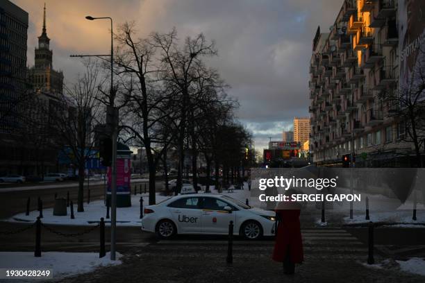 Pedestrians wait for a green traffic light to cross a road as a Uber car passes near the Palace of Culture snd Science in the background, on a sunny...