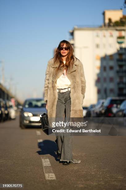 Estelle Chemouny wears sunglasses, a fluffy faux fur winter jacket, a white top, a belt, gray denim ripped flared jeans, black leather shoes, a...