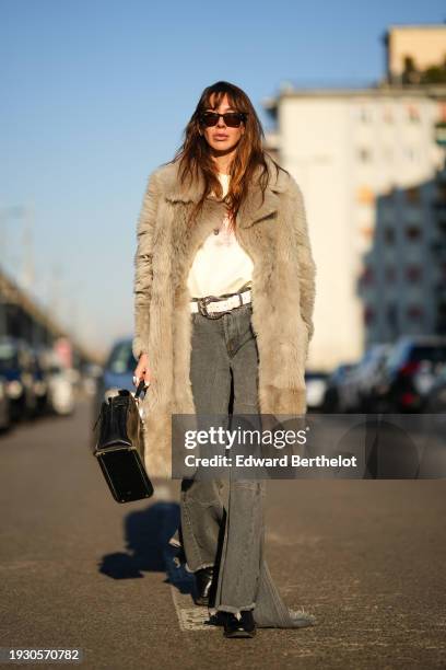 Estelle Chemouny wears sunglasses, a fluffy faux fur winter jacket, a white top, a belt, gray denim ripped flared jeans, black leather shoes, a...