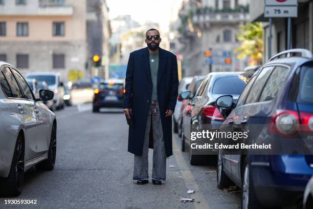 Jean-Claude Mpassy wears sunglasses, a pullover, a dark navy blue long coat, gray jeans denim pants, leather shoes, outside Dolce & Gabbana, during...