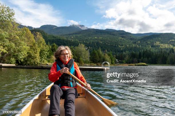 senior woman paddles canoe across lake - seniors canoeing stock pictures, royalty-free photos & images