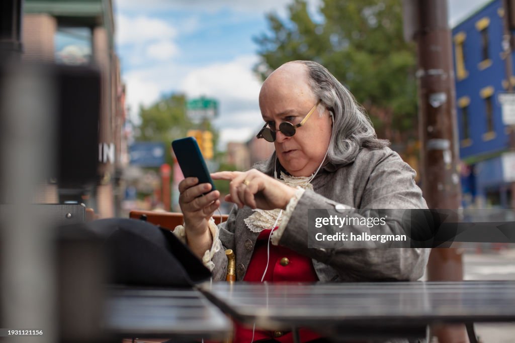 Benjamin Franklin Impersonator Tapping his Smartphone While Sitting in Outdoor Patio of Bar