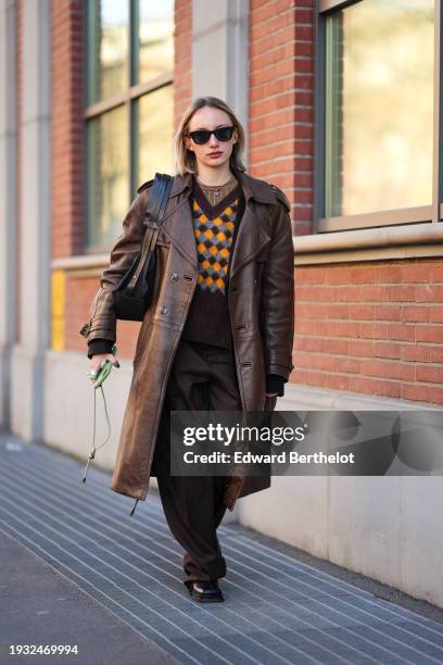 Guest wears sunglasses, a necklace, a v-neck brown orange and gray checkered pattern pullover, a brown long leather coat, oversize suit pants,...