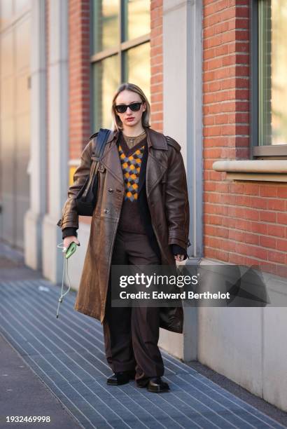 Guest wears sunglasses, a necklace, a v-neck brown orange and gray checkered pattern pullover, a brown long leather coat, oversize suit pants,...