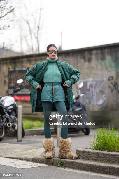 Guest wears silver mirror sunglasses, a green wool knit ribbed turtleneck pullover, a long bomber jacket, mini shorts, tights / leggings, brown...