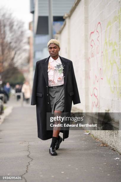 Guest wears a black long coat, a pale pastel pink shirt with floral details, a belt, gray shorts, black socks, black leather shiny pointed shoes,...