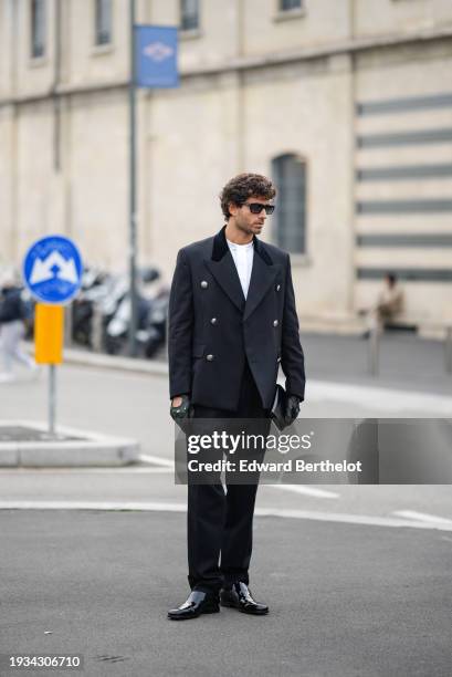 Guest wears a military / blazer jacket, a white t-shirt, black leather gloves, black pants, black shiny leather pointed shoes, outside Prada, during...