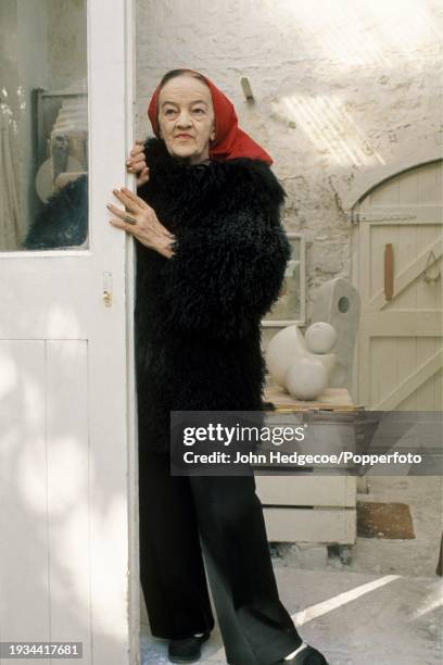 English artist and sculptor Barbara Hepworth posed wearing a thick wool coat and red headscarf at the door of her studio workspace, Trwyn Studios in...