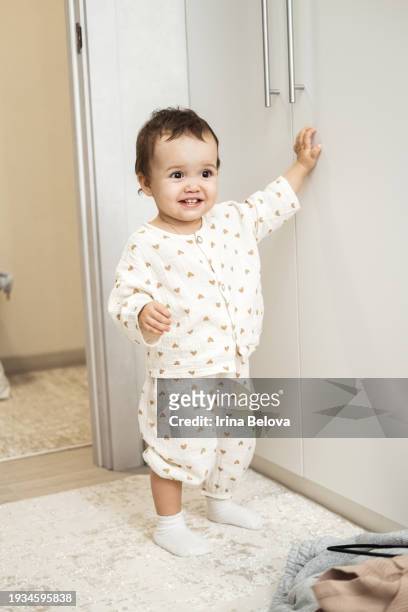 a happy kid in pajamas learns to walk, holds on to the wall with his hand and takes his first steps. - baby first steps stock pictures, royalty-free photos & images