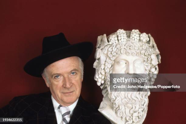 English fashion and portrait photographer and designer Sir Cecil Beaton posed standing next to a bust of an ancient Greek figure in England in 1974.