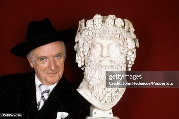 English fashion and portrait photographer and designer Sir Cecil Beaton posed standing next to a bust of an ancient Greek figure in England in 1974.