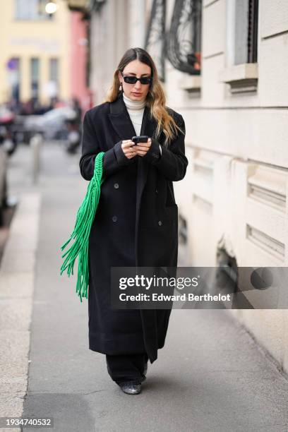 Guest wears sunglasses, earrings, a white turtleneck pullover, a black long coat, a green fringe bag, outside K-WAY, during the Milan Fashion Week -...